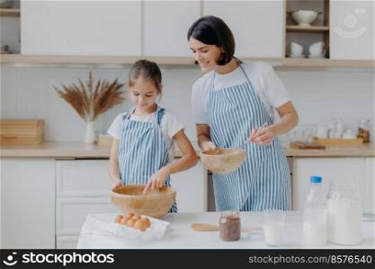 Preparing delicious meal and cooking together concept. Pleased mother looks attentively at how child whisks ingredients in bowl, wear aprons, stand in kitchen. Kid takes cooking lesson in mum
