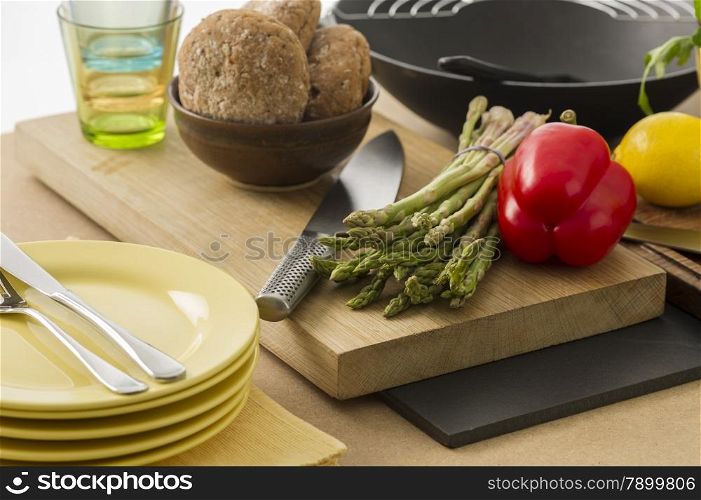 Preparing dinner with fresh vegetables. Preparing dinner with fresh vegetables in the kitchen with a bunch of green asparagus spears and a colorful red sweet bell pepper on a chopping board with plates and rolls