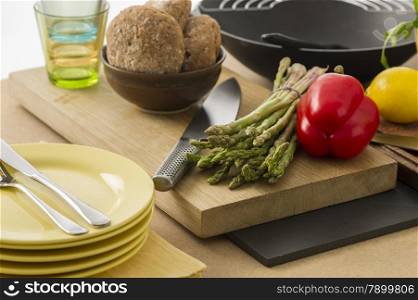 Preparing dinner with fresh vegetables. Preparing dinner with fresh vegetables in the kitchen with a bunch of green asparagus spears and a colorful red sweet bell pepper on a chopping board with plates and rolls