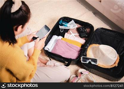 Preparing for travel in new normal. Woman chatting surfing internet use smartphone application during packing suitcase baggage, Female texting message on mobile phone with friend for ready