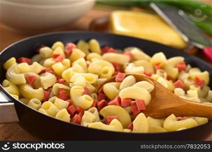 Preparing fresh homemade elbow macaroni pasta with sausage pieces in frying pan with cheese and green onion in the back (Selective Focus, Focus on the front of the food on the wooden spoon)