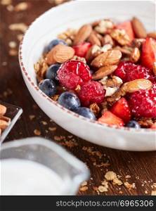 Preparing of natural breakfast with fresh organic ingredients - berries, granola, nuts, honey in a white bowl with milk on a wooden table.. Fresh berries, almonds, oatmeal and granola in a plate with milk on a wooden background. Healthy food