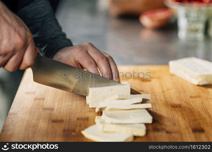 Preparing vegan meal. Chef&rsquo;s hand cutting tofu cheese with knife on wooden board. Chef&rsquo;s Hands Slicing Tofu Cheese with Knife on a Wooden Cutting Board