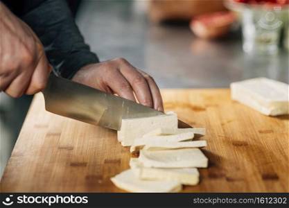 Preparing vegan meal. Chef&rsquo;s hand cutting tofu cheese with knife on wooden board. Chef&rsquo;s Hands Slicing Tofu Cheese with Knife on a Wooden Cutting Board