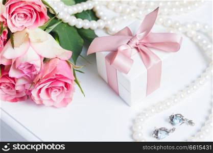 Present box with pink ribbon with jewellery and roses on white table. Box with pink ribbon