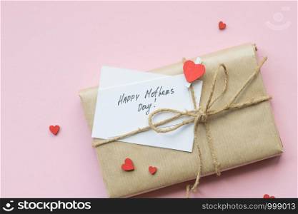 Present wrapped in brown craft paper and tie hemp string on Light pink background. Gift box with greetings on Mothers Day and decorative red hearts. Top view. Copyspace. Present wrapped in brown craft paper and tie hemp string on Light pink background. Gift box with greetings on Mothers Day. Top view.