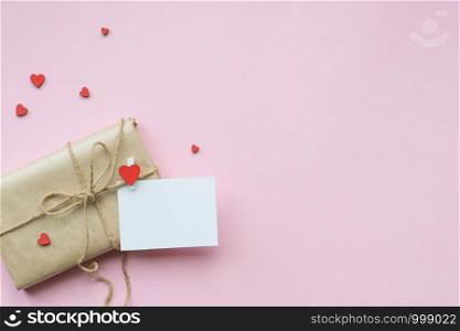 Present wrapped in brown craft paper and tie on Light pink background. Gift with decorative red hearts and empty greeting card for Mothers Day, Wedding, Birthday and Valentines Day. Top view. Copyspace. Mockup. Present wrapped in brown craft paper and tie hemp string on Light pink background. Romantic Gift with decorative red hearts. Top view.