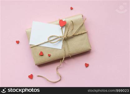 Present wrapped in brown craft paper and tie on Light pink background. Gift with decorative red hearts and empty greeting card for Mothers Day, Wedding, Birthday and Valentines Day. Top view. Copyspace. Mockup. Present wrapped in brown craft paper and tie hemp string on Light pink background. Romantic Gift with decorative red hearts. Top view.