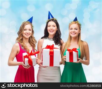 presents, holidays, people and celebration concept - smiling women in party caps with gift boxes over blue lights background
