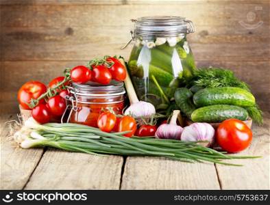 preserved tomatoes and cucumbers with fresh vegetables on wooden background