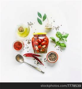 Preserved tomatoes in jar and preserving ingredients for flavoring: olive oil, spices, herbs, basil leaves, chili, garlic, pepper, bay leaves and spoon on white background. Top view.