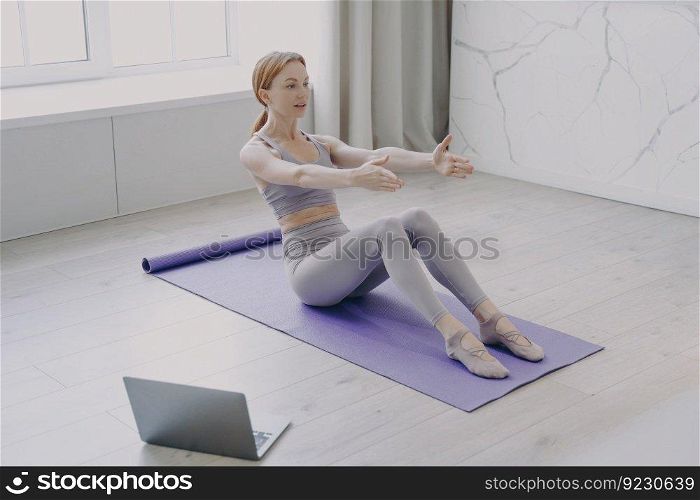 Press exercise and pilates at home. Slim sporty european girl is stretching on mat. Concept of distance learning and online fitness classes. Video lessons on laptop. Computer on floor.. Press exercise and pilates at home. Video lessons on laptop. Concept of distance fitness classes.