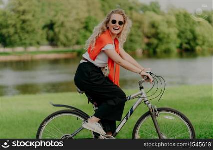Pretty active female bicyclist rides bike, looks at camera with smile, wears casual everyday clothes, poses near river, green trees, grass, spends free time in countryside. People, rest, lifestyle