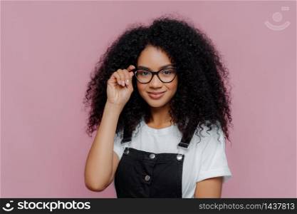 Pretty African American woman keeps hand on frame of glasses, smiles happily, wears white casual t shirt and black overalls, enjoys spare time, poses over violet background. Ethnicity, fashion concept