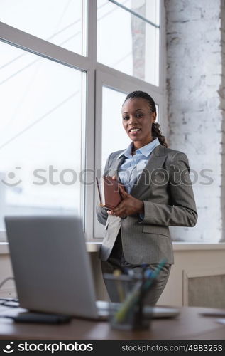 Pretty african business woman standing near window leaning windowsill at her office and taking notes in her diary while planning her day