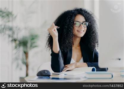Pretty Afro American woman looks positively at display of computer, writes down information from internet, wears optical glasses and black suit, sits at desktop with necessary things for work