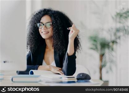 Pretty Afro American woman looks positively at display of computer, writes down information from internet, wears optical glasses and black suit, sits at desktop with necessary things for work