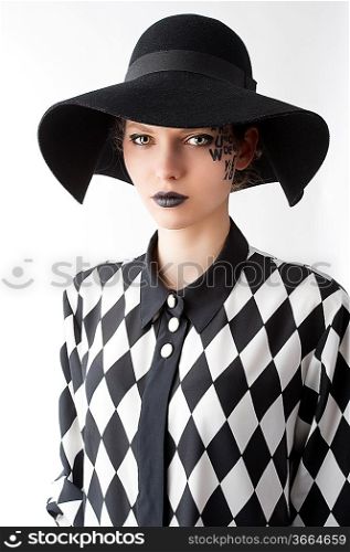 pretty and beautiful young lady with a creative make up made with letter on face and black hat and unusual shirt, she is in front of the camera and looks in to the lens with serious expression