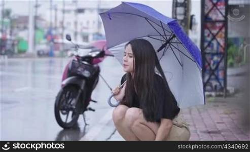 pretty asian girl holding umbrella sit down on the street side walk on raining day, rainy season reach her hand out touching rain drops, broke down motorbike, feeling disappointed stuck in the rain