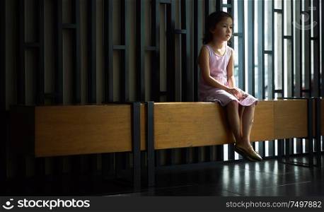 Pretty asian little girl sitting lonely on wood long chair at corridor .