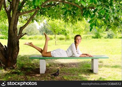Pretty barefooted woman is laying on a bench under the green tree. Black pigeon is walking around her.