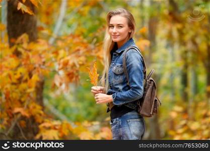 Pretty blond girl standing with beautiful dry maple leaf in hand in the autumn forest, enjoying beauty of fall nature, spending weekend in the park