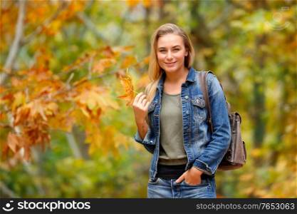 Pretty blond girl standing with beautiful dry maple leaf in hand in autumn forest, enjoyng beauty of fall weather, spending weekend in the park