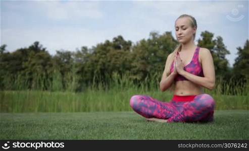 Pretty blond woman barefoot making yoga meditation in lotus pose on green grass on the park lawn