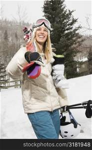 Pretty blond young woman carrying ski equipment in snow and smiling.