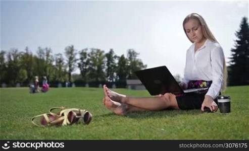 Pretty blonde businesswoman sitting on the green grass barefoot on the lawn in the park and answering a phone call while working on laptop