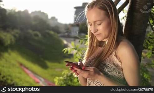 Pretty blonde female in fashionable dress texting message on mobile phone against urbanscape background while leaning on the tree in the park at sunset. Smiling girl browsing the net with smartphone outdoors. Slow motion. Steadicam stabilized shot.