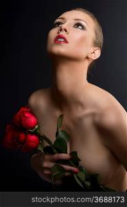 pretty blonde girl&acute;s low key portrait with three red roses, she is turned at three quarters at right, looks up and takes the roses near the chest