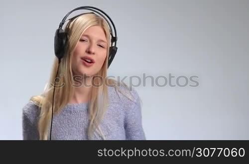 Pretty blonde girl having fun and listening to music in headphones over white background. Young attractive woman wearing modern headphones enjoying her favorite song, singing, subtly swaying along with music, moving groovily to the beat.