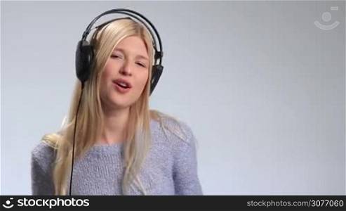 Pretty blonde girl having fun and listening to music in headphones over white background. Young attractive woman wearing modern headphones enjoying her favorite song, singing, subtly swaying along with music, moving groovily to the beat.