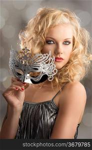 pretty blonde girl with curly hair takes one silver mask, her body is turned of three quarters at left , she looks in to the lens and takes the mask with right hand