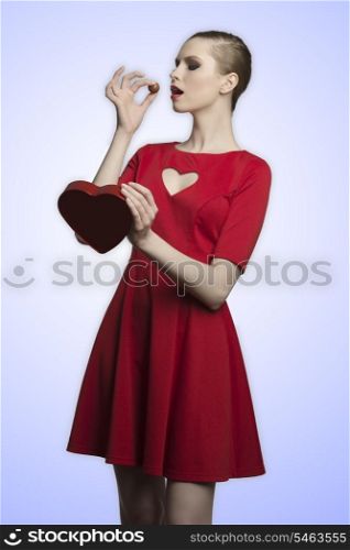 pretty blonde girl with red romantic dress and cute make-up eating one praline from the heart shaped box in her hands. Valentine?s day