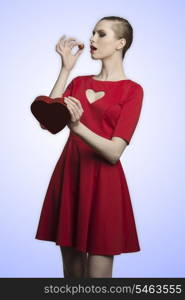 pretty blonde girl with red romantic dress and cute make-up eating one praline from the heart shaped box in her hands. Valentine?s day