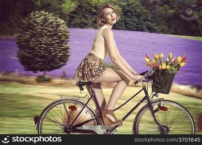 pretty blonde woman going on the bicycle with colourful flowers in the basket. She wearing pink tank and very short floral skirt. rural field lavander