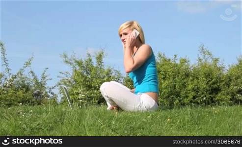 Pretty blonde woman seated on a lush green lawn with her laptop in front of her talking on her mobile phone