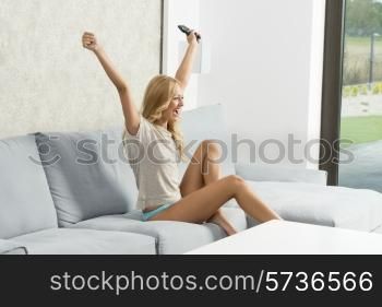 pretty blonde woman sitting with naked legs and panties on sofa, exulting with happiness expression watching television and taking remote control in the hand