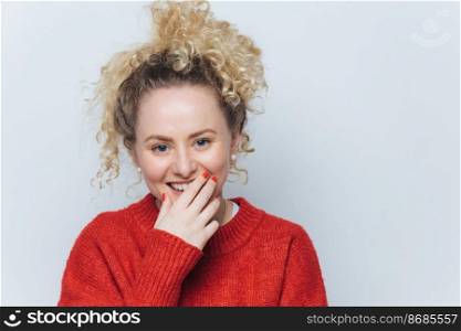 Pretty blue eyed female with cheerful expression, giggles and covers mouth with hand, expresses happiness, dressed casually, isolated over white studio background. Lovely blonde curly woman.