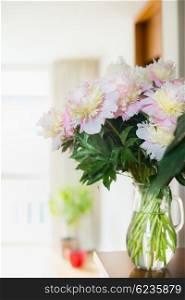 Pretty Bouquet of pink pale peonies in a glass jug with water on the table against the background of a bright room. Home decoration and interior