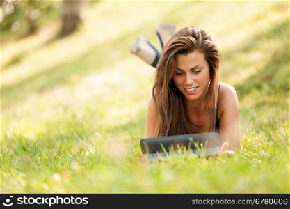 Pretty brunette girl is looking a digital screen while sitting on the grass and smiling