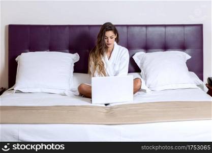 Pretty brunette in white bathrobe sitting on bed in hotel room and working on laptop in relax. Woman working on laptop in hotel