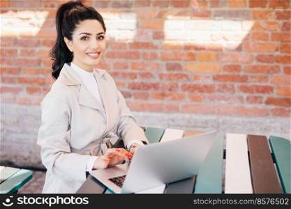Pretty brunette student female wearing elegant white coat working at computer writing her thesis looking with smile at camera. Businesswoman in elegant clothes sitting indoors over brick wall