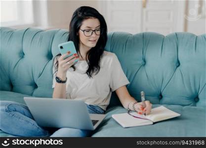 Pretty brunette student watches webinar, takes notes on sofa, holds phone, busy with distance job. People, technology, lifestyle concept.