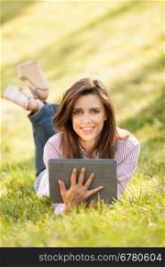 pretty brunette woman laying on a grass field while looking on a digital tablet