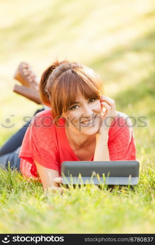 pretty brunette woman laying on a grass field while looking on a digital tablet