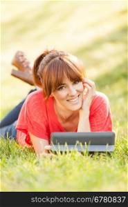 pretty brunette woman laying on a grass field while looking on a digital tablet