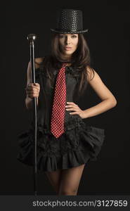 pretty brunette woman wearing like cabaret actress with black corset, red tie and hat taking stick in the hand on black background